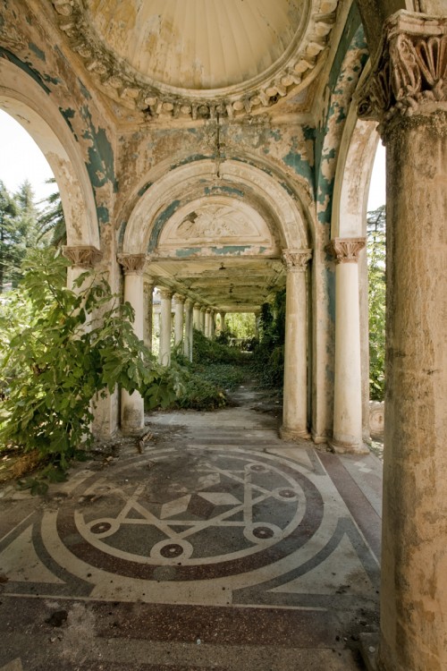 A photo of a walkway in an abandoned railway station. The architecture is ornate but deteriorating, and plants have started to overgrow parts of the structure.