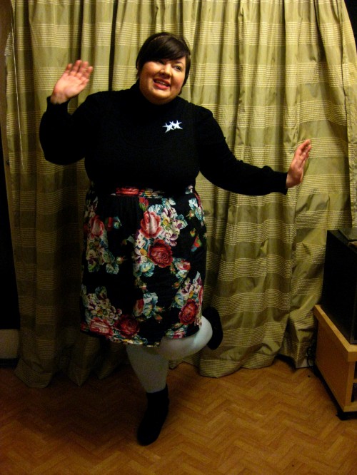 Photo of Natalie in a black turtleneck and floral skirted dress with light mint stockings and black boots, posing facetiously in a dance-like stance.