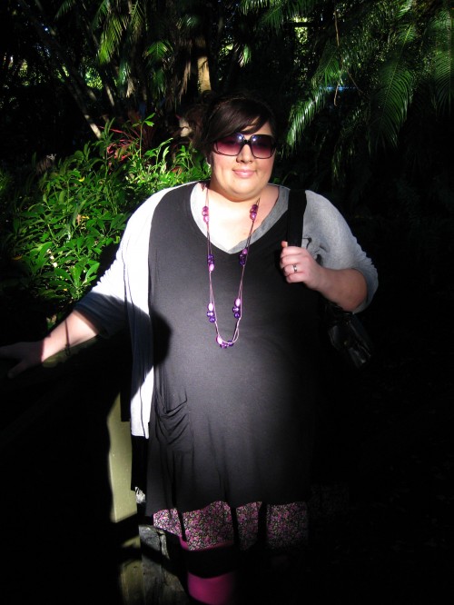 Photo of Natalie in patchy light of a rainforest type setting wearing long black top with  a grey cardigan, long purple necklace and sunglasses.