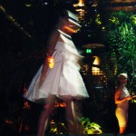 A model wears a short white dress with a full skirt and a large bow at the bust.
