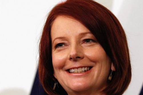 Close up photo of the face of Julia Gillard, 27th Prime Minister of Australia, a white woman with chin length red hair. She is grinning. A small part of the Australian flag is visible behind her.