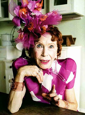 A photo of an older woman dressed in fucshia sits in a kitchen, bright pink and orange flowers in her red hair, her right hand brought up under her chin and her left hand gesturing towards the camera.
