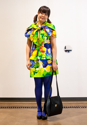A street style photo of a young woman wearing a brightly printed dress of acid green, yellow, purple and blue wearing blue tights and mary jane style shoes, carrying a big black shoulder bag.