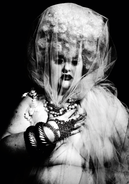 A black and white photo of a pale skinned plus size model wearing a curly blonde wig with tulle pulled over her head. Her hand clutches at the tulle on her chest and she's wearing jewellery draped over her hand with big bangles.