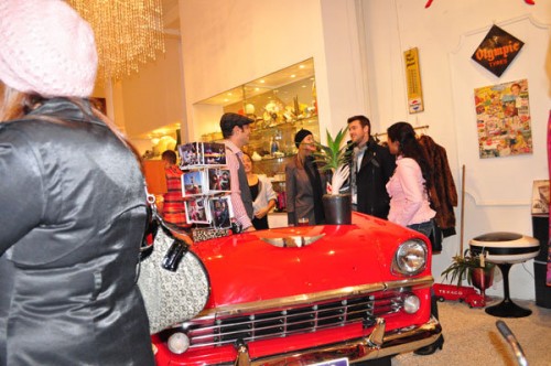 Photo of a shop with the hood of a red car in the center of the room, people crowded around talking to each other.