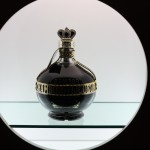A bottle of Chambor (a dark purple liqeur in a gold accented fancy bottle) sits on a shelf with a white background.