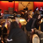 A crowded bar with low grey/ fawn couches with a long bar in the background. Behind the bar is red curtains and a pair of antlers on the wall.