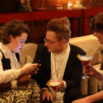 A woman on her phone, a man holding a martini and another woman holding a martini chat.