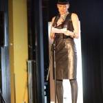 A woman stands behind a microphone reading from prompt cards. She wears a black leather dress, black leggings and black sandals.