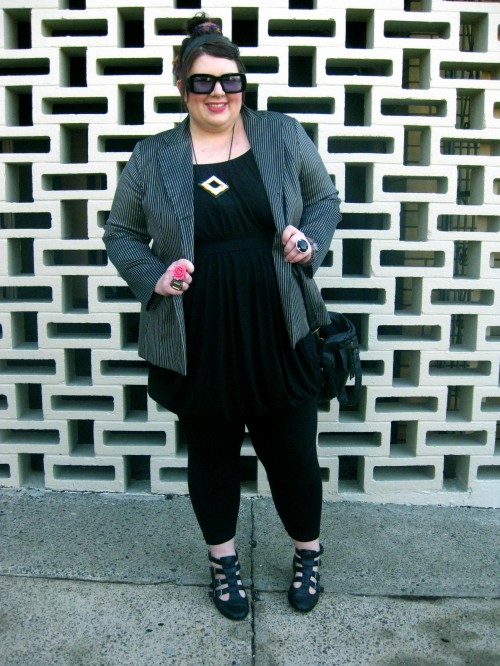 A photograph of Natalie against a decorative brick wall. She wears a black bubble dress over black leggings with a gold/black pin striped blazer and sandal boots with buckles up the front.