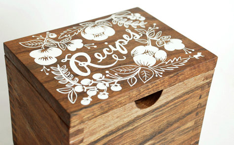 A small wooden recipe card box with a white screenprinted floral design on the lid and handwritten lettering saying "Recipes".