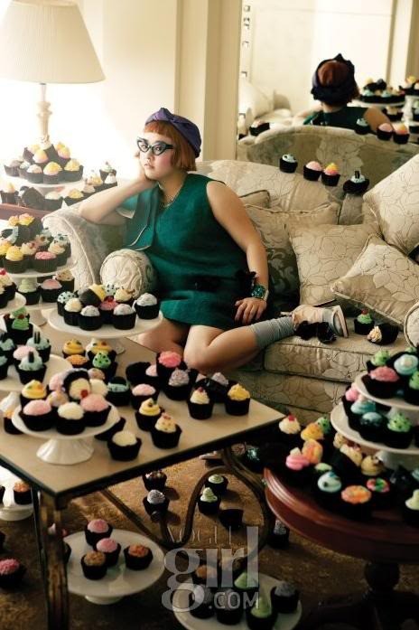 A young woman sits on a couch, surrounded by hundreds of brightly frosted cupcakes.
