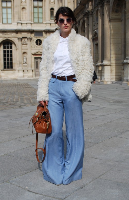 A young slim pale skinned woman wearing a white buttoned up shirt with blue flared jeans and a brown belt. Over the outfit she wears a white fur (looks faux!) jacket, and she carries a tan leather bag.