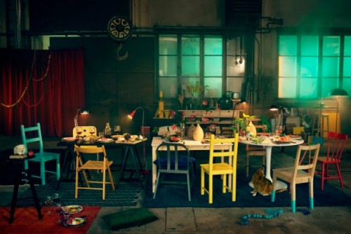 A photo of a warehouse style room with mismatched chairs and tables set up for a dinner party. Food, desklamps, wine glasses and dinner plates clutter the tables, some are even on the floor.