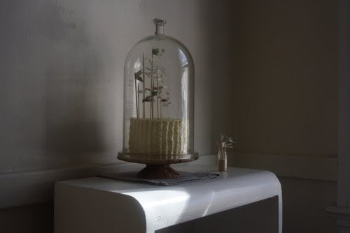 A cake with many little cake flags, all of different heights, sits inside a tall cake "jar" (I guess!) on a white bench.