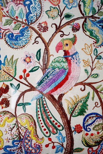 Detail photo of an embroidered piece of fabric with a brightly coloured bird sitting in a tree with paisley-like symbols as the foliage.
