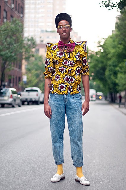 A street style photo of a young slim brown skinned man wearing a bright yellow print shirt with pointy shoulders, big pink bow tie, pegged and too short stone wash jeans with yellow socks and cream loafers. He also wears a cap tilted back on his head and yellow rimmed glasses.