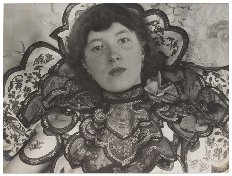 Black and white photo of a young pale skinned woman with chin length hair wearing an elaborately layered and embroidered neckpiece with scalloped edges. A rose sits on her left shoulder, she stares at the camera with an unsmilling, but not unkind, face