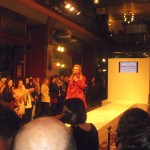 A woman with long blonde hair in a red jacket stands at the end of a white runway with a microphone, people crowded around.