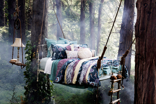 A photo of a bed with a handmade quilt and lots of cushions that is suspended by rope from trees in the middle of a forest.