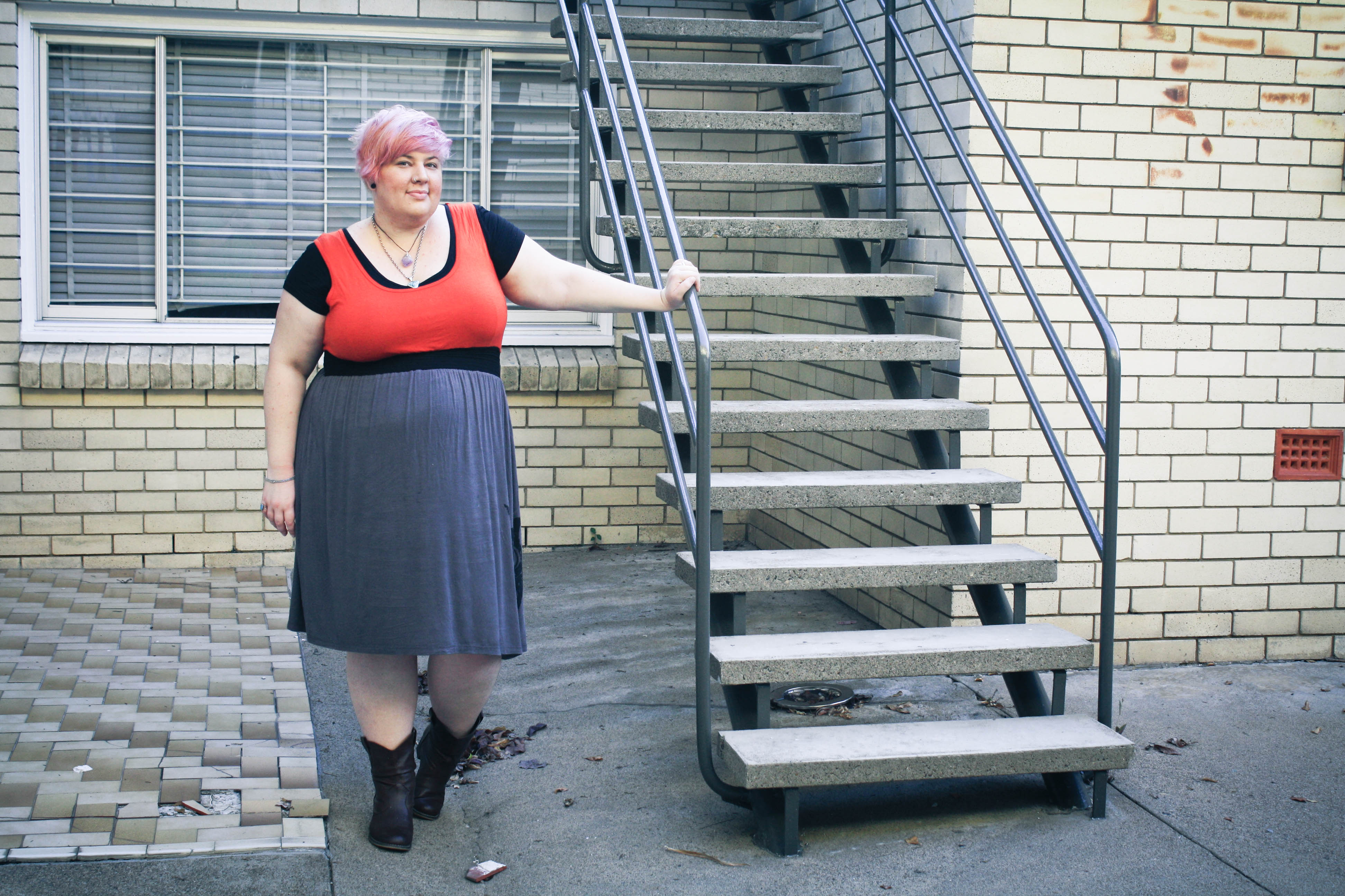 Photo of me wearing a dress with a red top and grey bottom with a black tshirt underneath and cowboy boots. I'm leaning against a stairway rail.