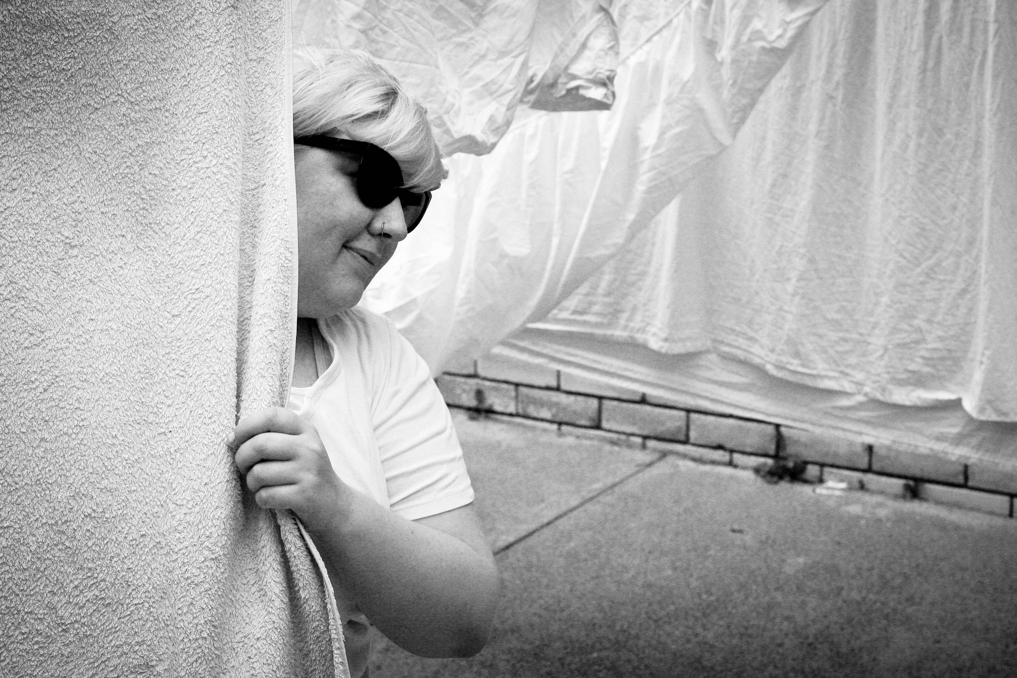 A black and white photo of me wearing sunglasses and peering around a towel on the line.