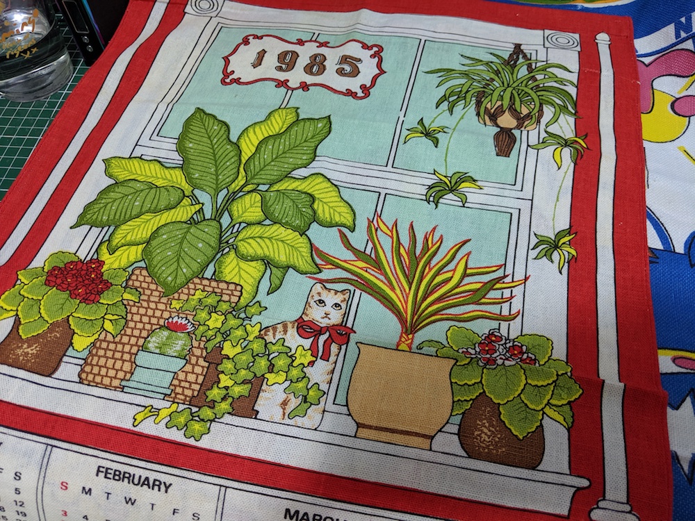 Photo of a tea towel with a lovely scene of house plants and a cat with a 1985 calendar on it.