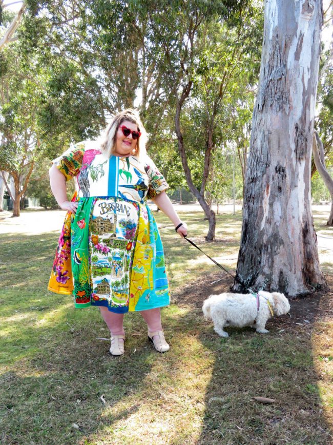 A photo of Natalie in the park wearing a brightly coloured dress made out of various Australian tea towels. She is holding Miffy on a leash. 